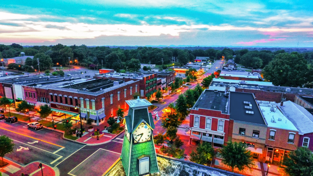 Aerial view of downtown Statesville NC at sunset, looking from the historic clock tower down Broad Street with lit-up shops and tree-lined sidewalks