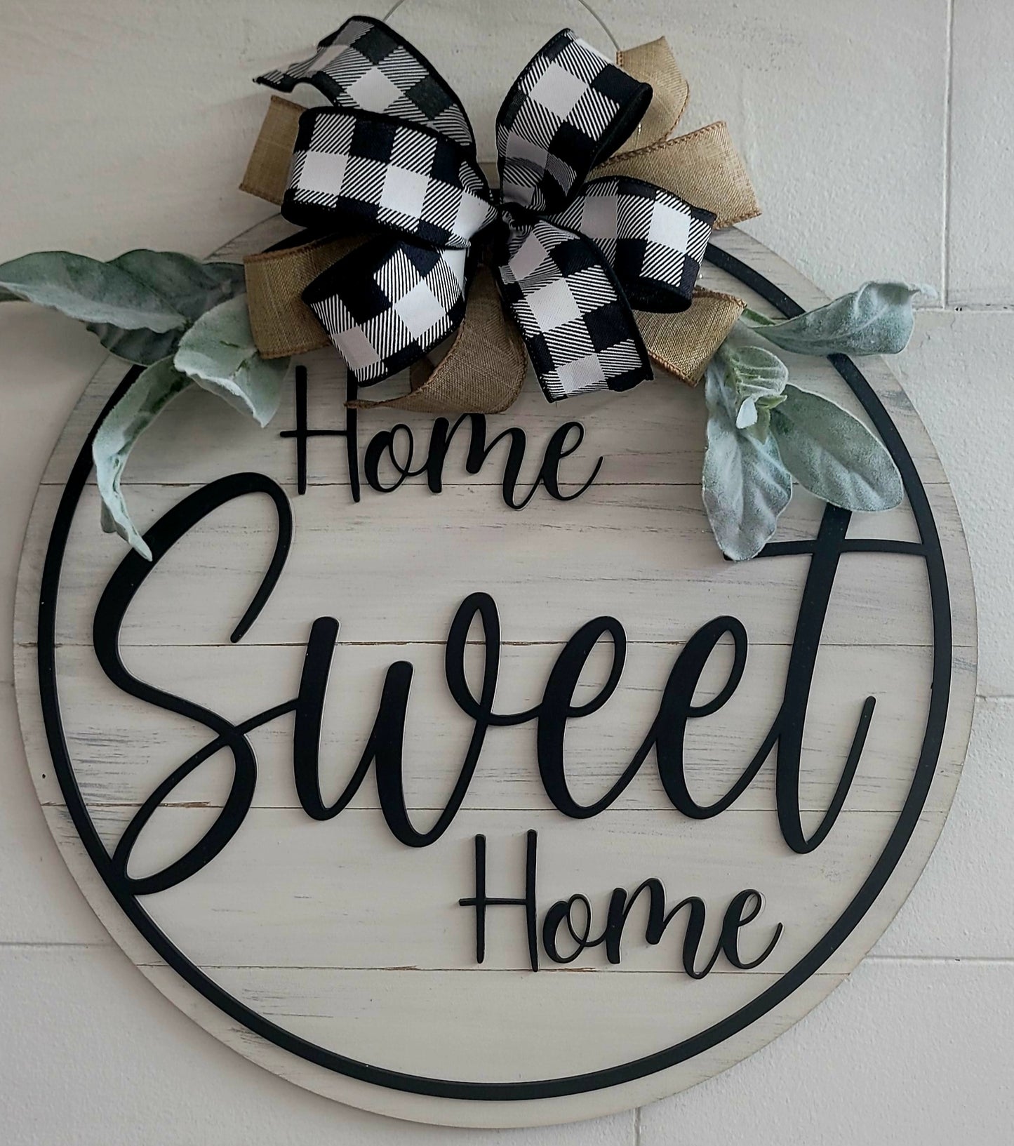 Rustic farmhouse "Home Sweet Home" round wooden door hanger sign with black script text, buffalo plaid and burlap bow, lamb’s ear greenery, and whitewashed wood background