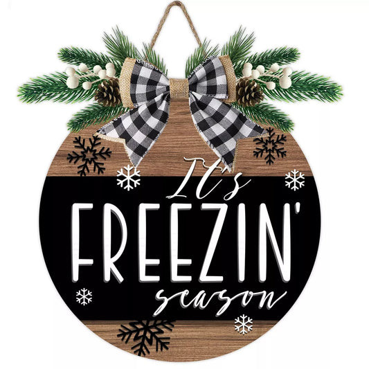 A circular door hanger with a winter theme, featuring text 'It's Freezin' Season' surrounded by snowflakes, evergreen branches, and pinecones.