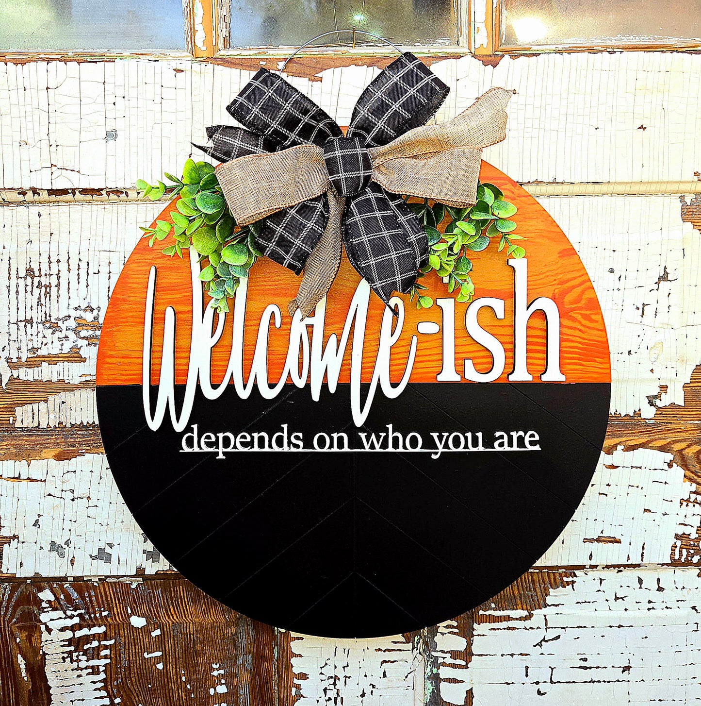 Humorous “Welcome-ish depends on who you are” round wooden door hanger featuring rustic orange and black two-tone background, playful white script, faux greenery, and a black-and-tan plaid bow – perfect for front porch decor, housewarming gift, or unique welcome sign.