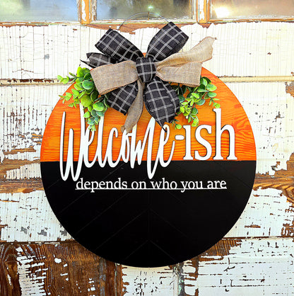 Humorous “Welcome-ish depends on who you are” round wooden door hanger featuring rustic orange and black two-tone background, playful white script, faux greenery, and a black-and-tan plaid bow – perfect for front porch decor, housewarming gift, or unique welcome sign.