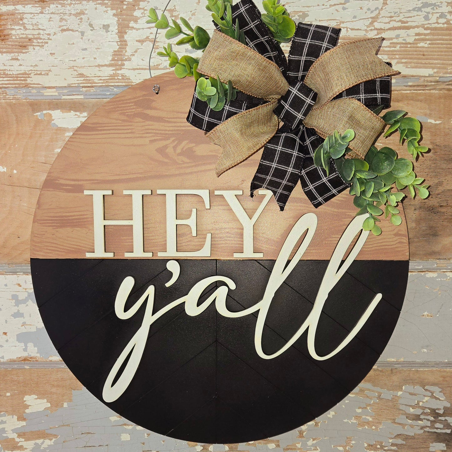 Rustic farmhouse-style “Hey y’all” round wooden door hanger with a two-tone natural wood and black finish, featuring bold cream lettering, black plaid and burlap ribbon bow, and faux greenery – perfect Southern welcome sign for front porch decor, entryway charm, or handmade gift