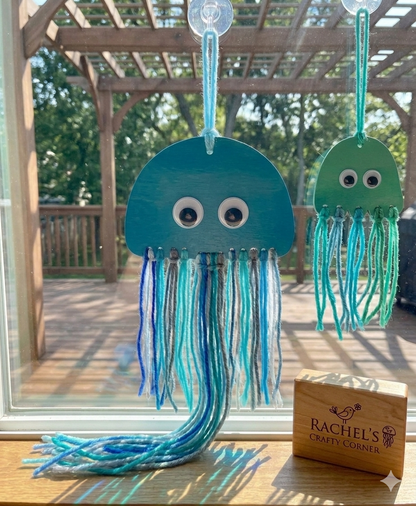 Two jellyfish-shaped decorations with string tails on a wooden surface, with 'Rachel's Crafty Corner' sign in the background.
