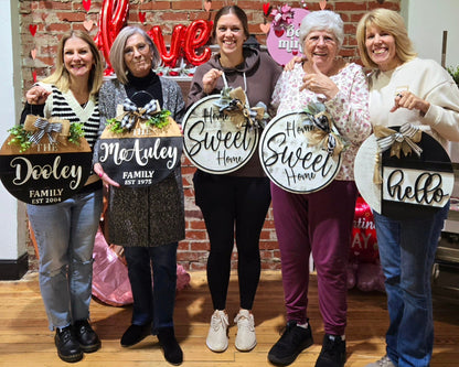 Happy group of women displaying custom wooden door hangers at a DIY craft workshop, featuring personalized family name signs and farmhouse-style designs with “Home Sweet Home” and “hello”