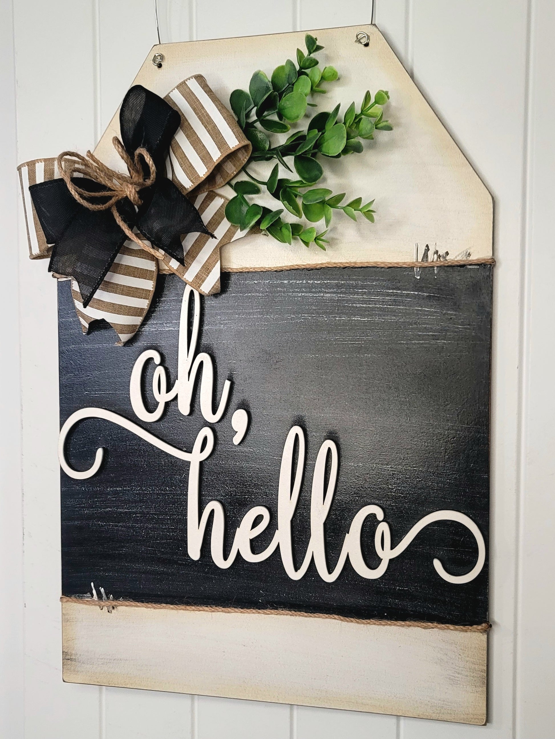 Rustic farmhouse “Oh, Hello” wooden door hanger sign with black and white script, striped burlap and black ribbon bow, faux greenery, and distressed wood finish – charming front porch or entryway welcome décor.
