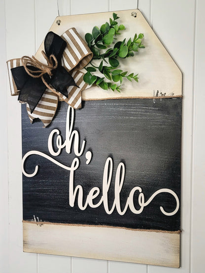 Rustic farmhouse “Oh, Hello” wooden door hanger sign with black and white script, striped burlap and black ribbon bow, faux greenery, and distressed wood finish – charming front porch or entryway welcome décor.